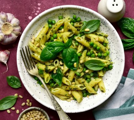 Penne cu sos pesto și mazăre într-o farfurie albă cu furculiță, alături de o solniță, o căpățână de usturoi, un bol cu semințe de pin și frunze de busuioc